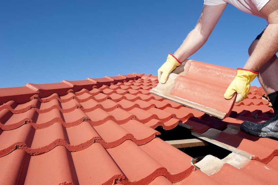 Roof repair, worker with yellow gloves replacing red tiles or shingles on house with blue sky as background and copy space; how long does it take to replace a roof