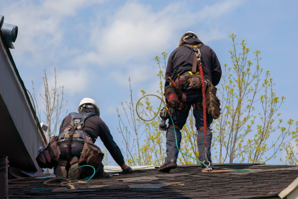 Two construction workers in safety gear and helmets are working on a rooftop under a clear sky, perhaps pondering how long does a roof last. One is crouching while the other stands, both using tools and safety harnesses. Young trees with budding leaves are visible in the background.